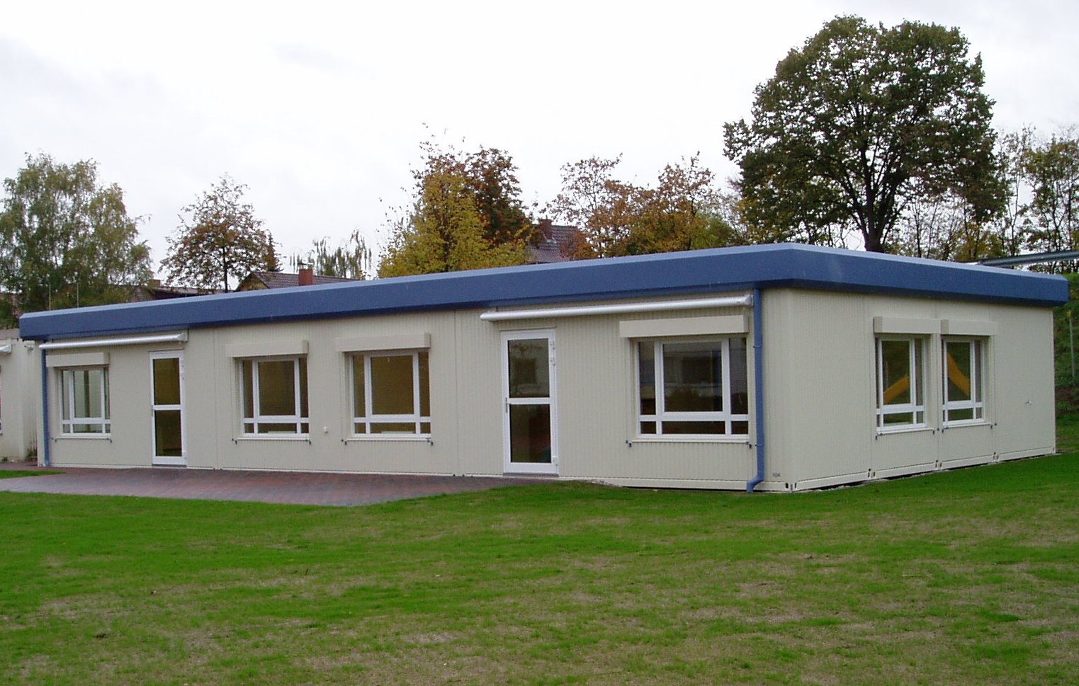 Gotowy modułowy budynek parterowy z płaskim, niebieskim dachem, beżową elewacją z kilkoma oknami z roletami i drzwiami, otoczony zielonym trawnikiem i drzewami w tle.