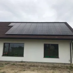 Panele fotowoltaiczne 9,9kWp zamontowane na brązowym dachu domu jednorodzinnego z białymi ścianami i dwoma oknami, widok z ziemi.