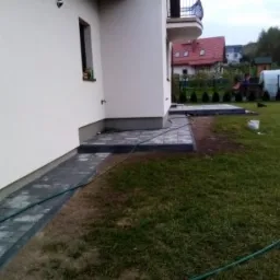 Ułożona ścieżka z kostki brukowej przy ścianie domu, z widocznym fragmentem trawnika i węża ogrodowego, teren jeszcze niezagospodarowany.