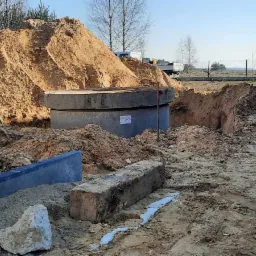 Betonowy krąg studni w wykopie, otoczony stertami piasku i elementami obrzeży, z widocznym fragmentem koparki i ciężarówki w tle.