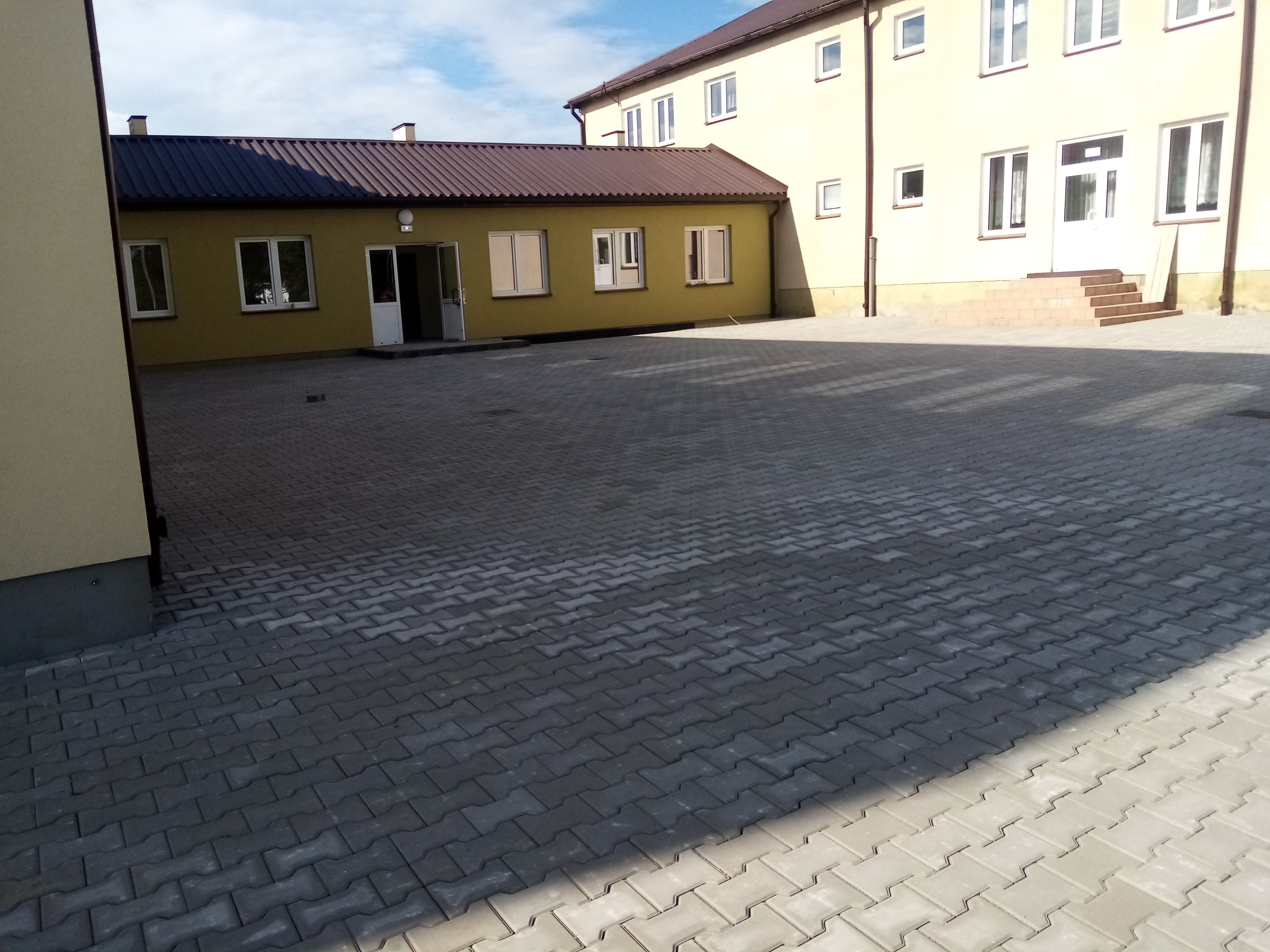 Szara kostka brukowa ułożona na dziedzińcu przed budynkiem, widoczny cień rzucany przez budynek, w tle budynek o żółtej elewacji.