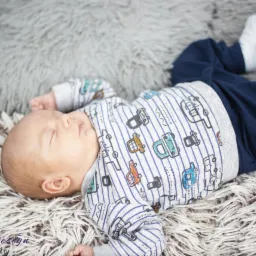 Śpiący noworodek ubrany w bluzkę w paski i autka leży na puszystym, szarym dywanie, z widocznymi białymi skarpetkami.