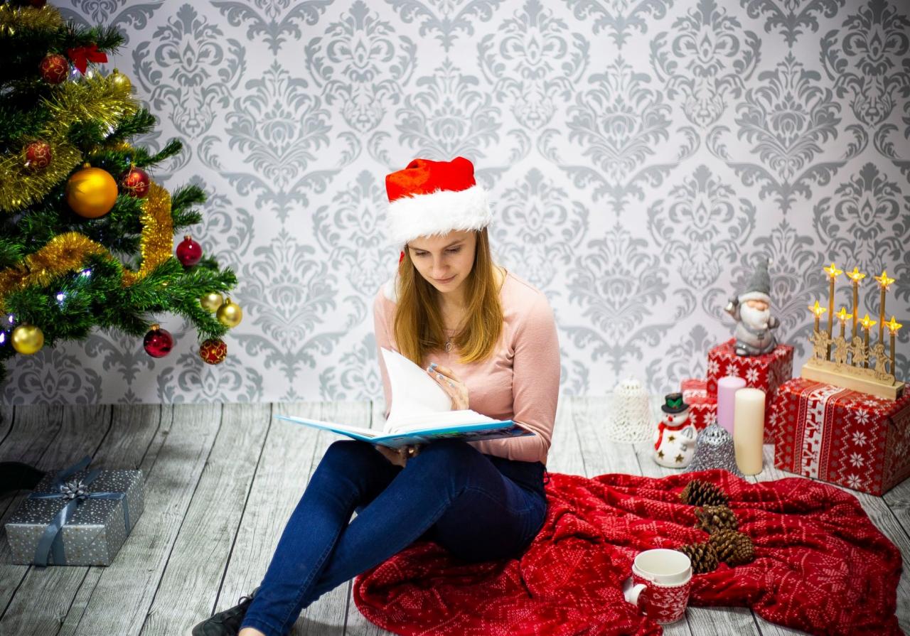 Kobieta w czapce Mikołaja czyta książkę siedząc na czerwonym kocu obok choinki, prezentów i dekoracji świątecznych na tle wzorzystej tapety.
