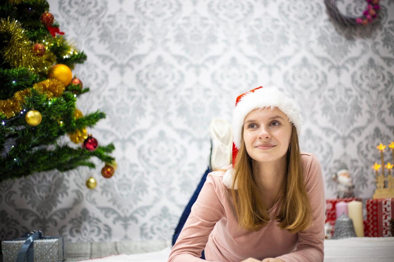 Uśmiechnięta młoda kobieta w czapce Mikołaja leży na białym futrze obok choinki, prezenty w ozdobnych papierach i świecznik w kształcie gwiazdy, tło stanowi ściana w ozdobne wzory.