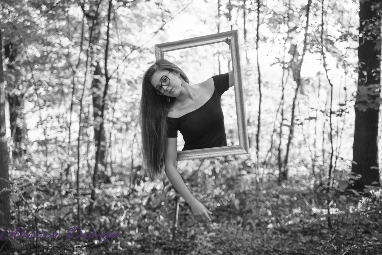 Czarno-biała sesja portretowa kobiety z okularami, trzymającej ramę obrazu na tle leśnym, z efektem wizualnym częściowego zniknięcia za ramą.