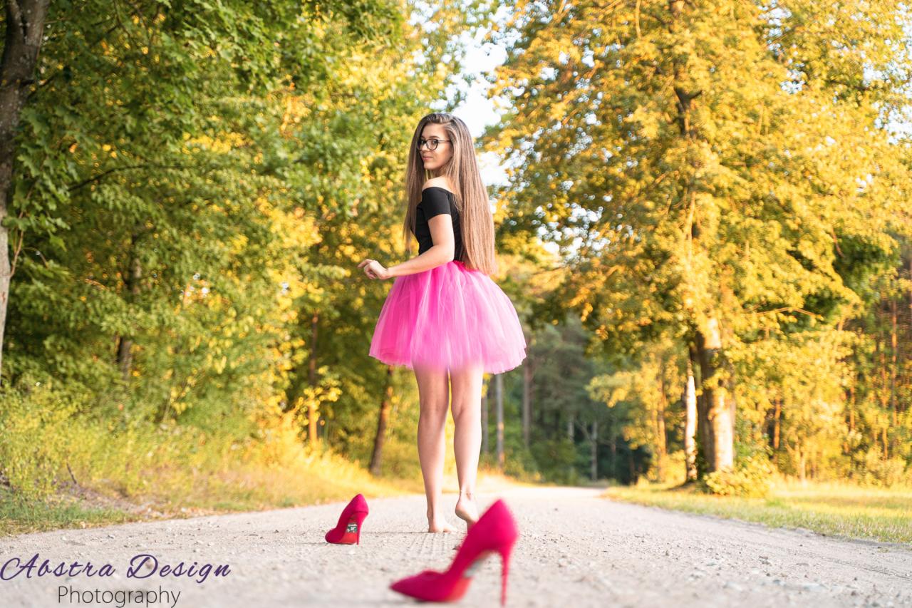 Młoda kobieta w różowej tiulowej spódnicy i czarnej bluzce, stojąca boso na leśnej drodze, z czerwoną szpilką leżącą obok, odwrócona plecami do aparatu, spoglądająca przez ramię, z rozpuszczonymi...