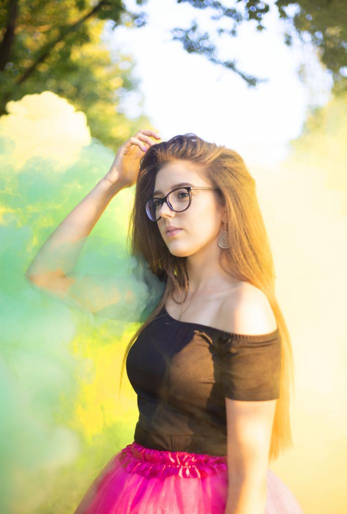 Młoda kobieta w okularach i czarnej bluzce z odkrytymi ramionami, różowa tiulowa spódnica, pozuje na tle zielono-żółtego dymu.