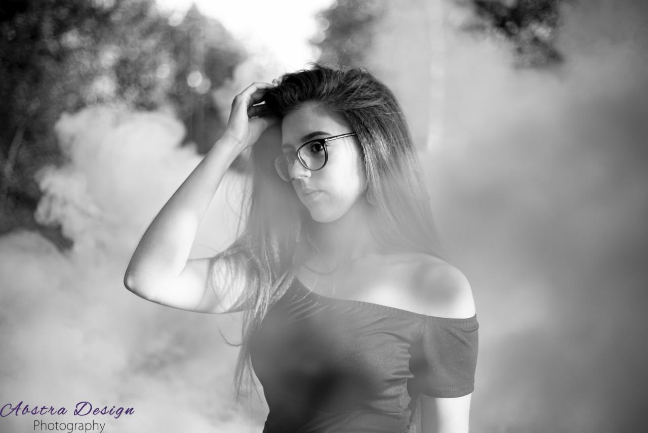 Portret młodej kobiety w okularach, z ręką we włosach, na rozmytym tle z efektem dymu, fotografia czarno-biała.