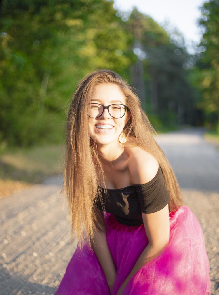 Uśmiechnięta młoda kobieta w okularach, czarnym topie i różowej tiulowej spódnicy pozuje na leśnej ścieżce w promieniach zachodzącego słońca.