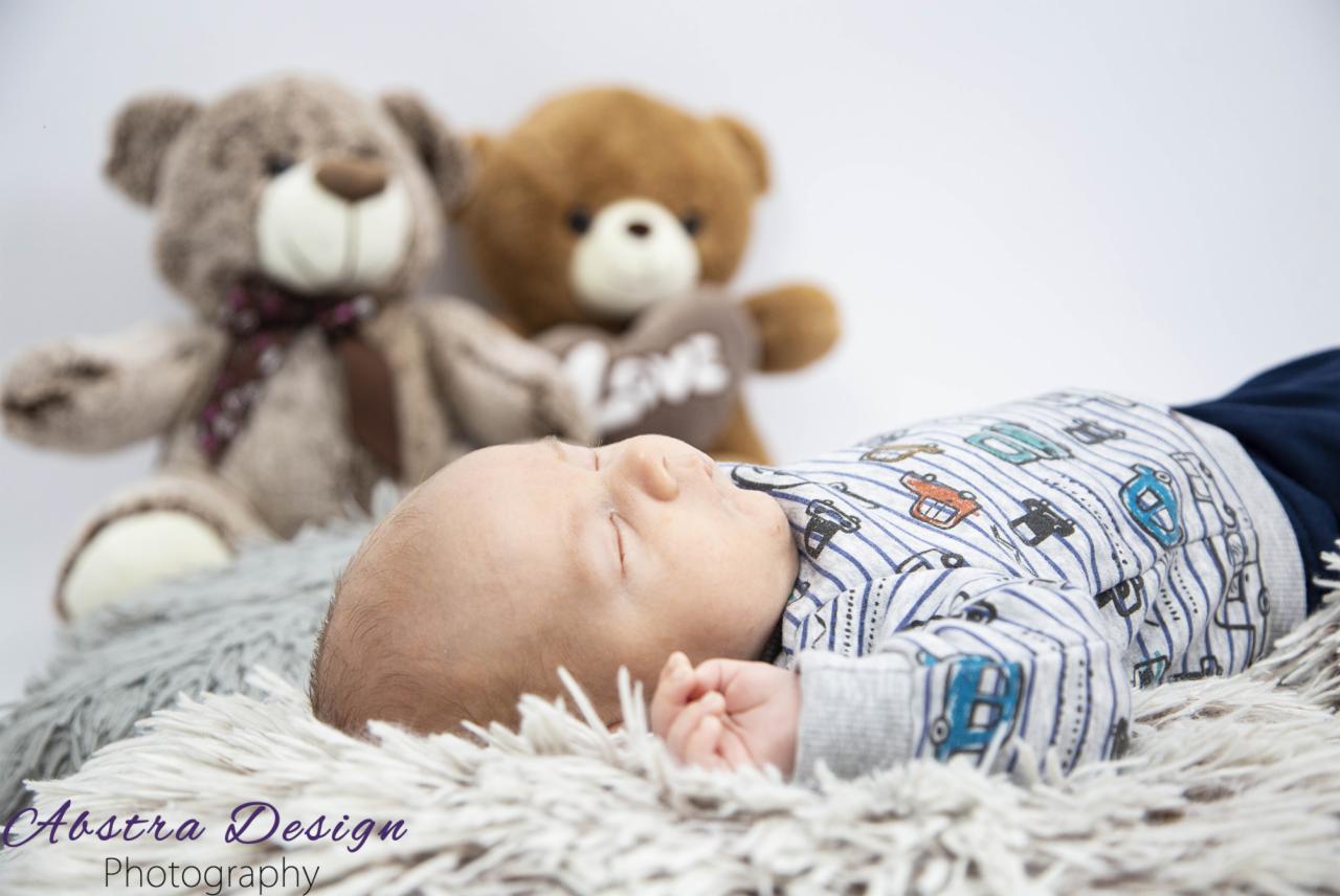 Śpiący noworodek w koszulce w paski z motywem samochodów, leżący na puszystym, jasnym dywaniku, w tle dwa pluszowe misie, jeden z napisem 'LOVE'.