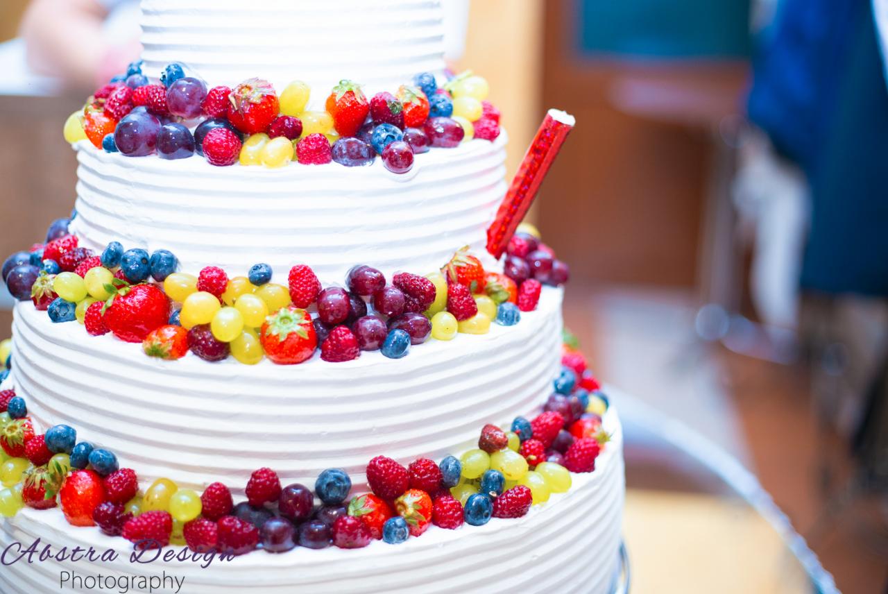 Trzypiętrowy tort weselny z białym kremem, udekorowany świeżymi truskawkami, malinami, winogronami i borówkami, z czerwoną świeczką w górnej warstwie.