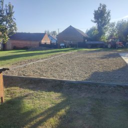 Kacper Kłosowski - Przygotowany teren pod trawnik z obrzeżami, widoczny traktor z przyczepą w tle, domy z czerwonej cegły i drzewa w otoczeniu.