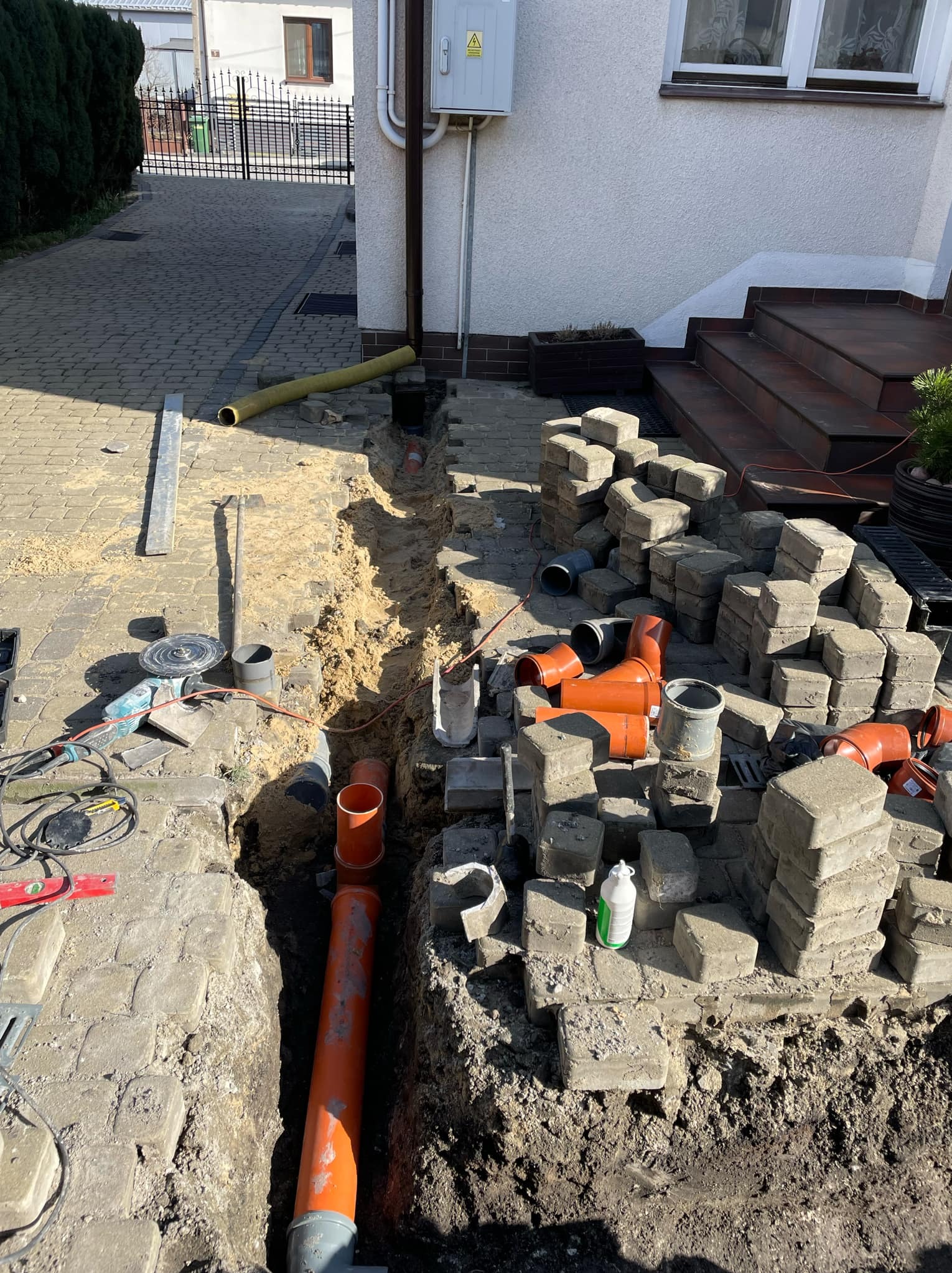 Wykop pod instalacje sanitarne w Zakrzówku: widok na wykop z ułożonymi rurami kanalizacyjnymi, kostką brukową i narzędziami do cięcia.