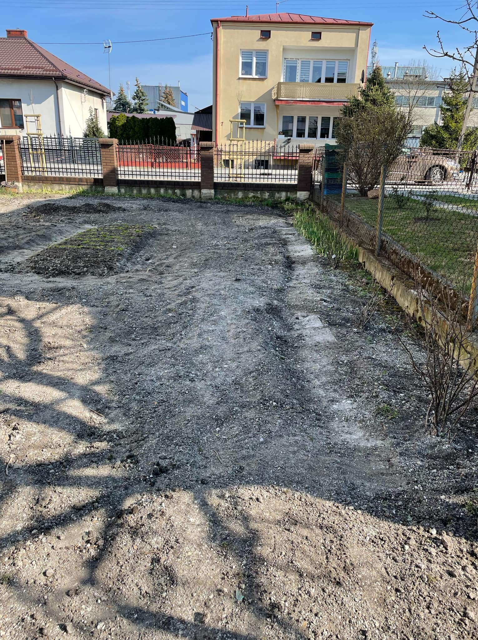 Przygotowany teren pod przyszły ogród, widoczne ślady po maszynach, częściowo wyrównana ziemia z niewielkimi kępkami trawy, otoczenie stanowią budynki mieszkalne i ogrodzenie.