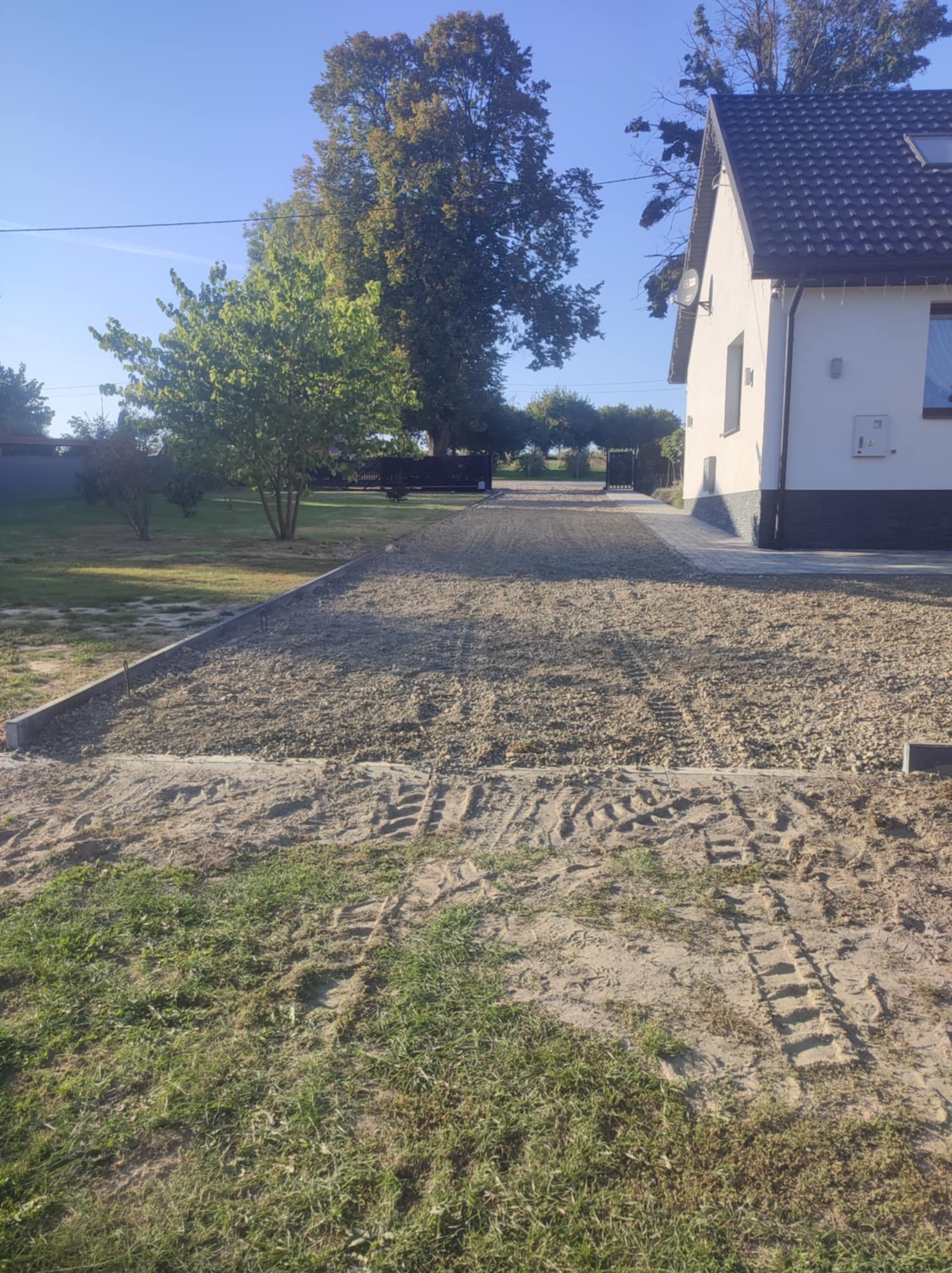 Przygotowany teren pod budowę podjazdu z obrzeżami betonowymi, widoczne ślady opon na piasku, dom jednorodzinny w tle.