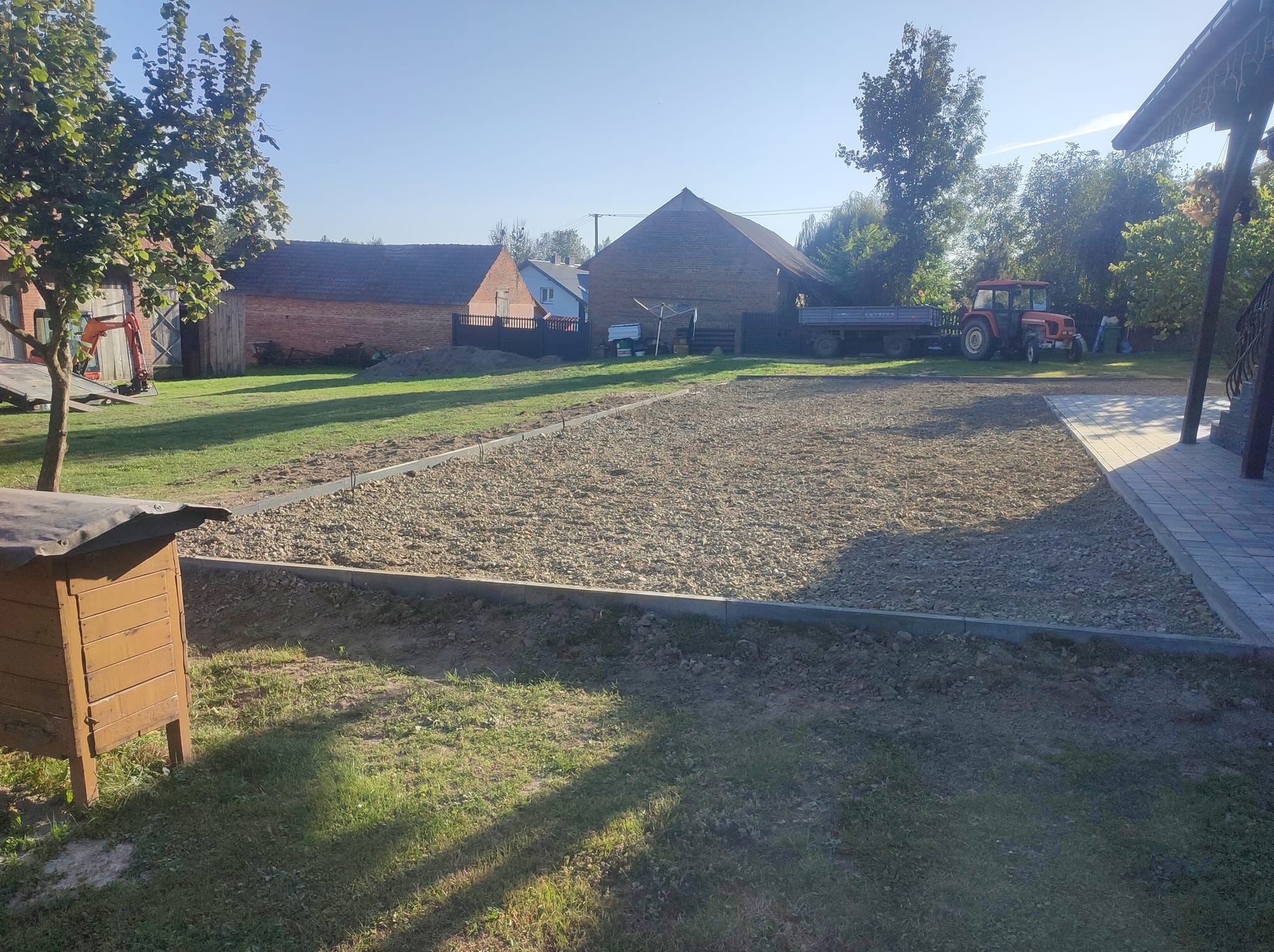 Przygotowany teren pod trawnik z obrzeżami, widoczny traktor z przyczepą w tle, domy z czerwonej cegły i drzewa w otoczeniu.