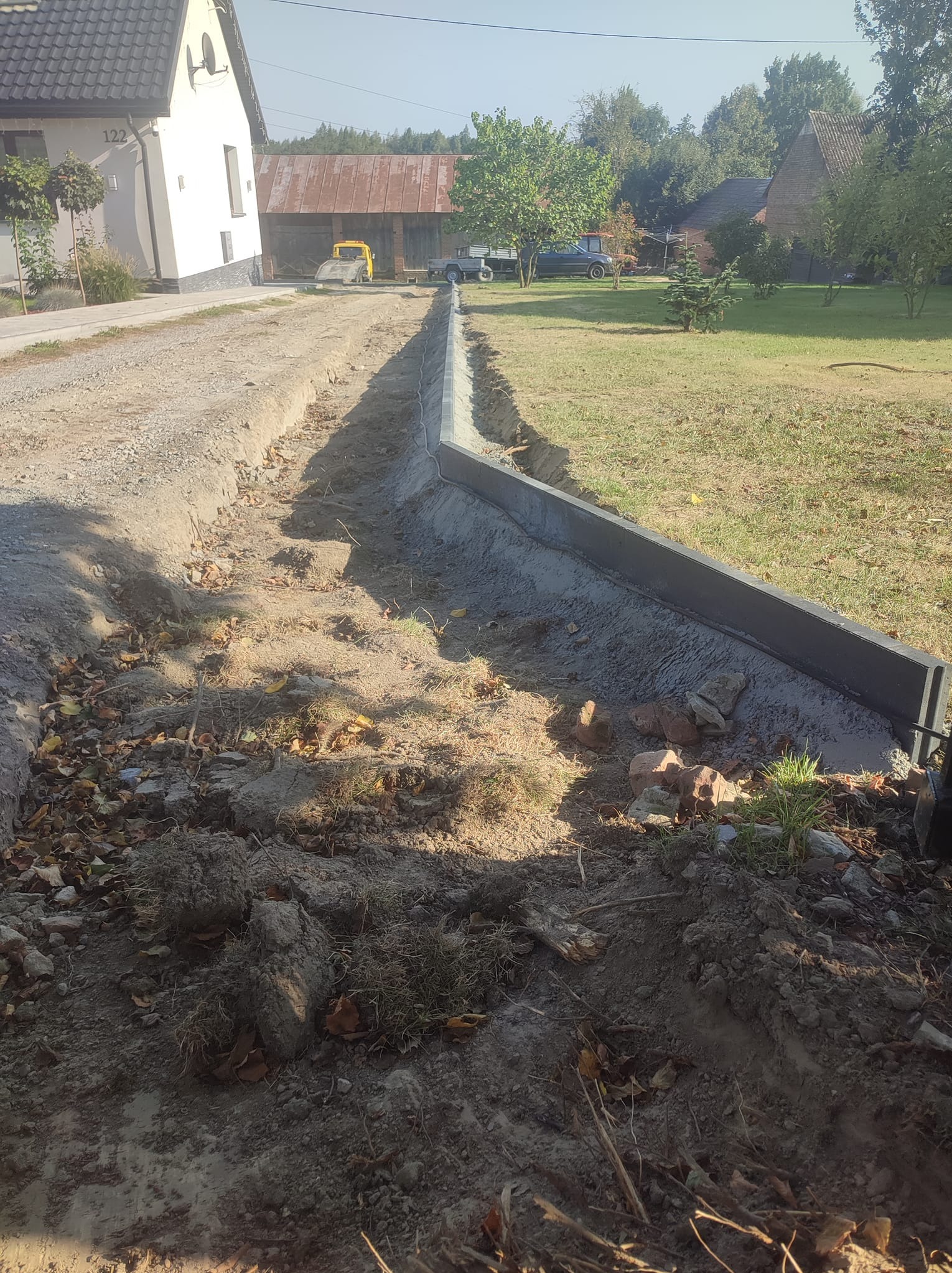 Świeżo wylany betonowy obrzeże w wykopie, widoczne ślady prac ziemnych, w tle dom z numerem 122 i zabudowania gospodarcze.