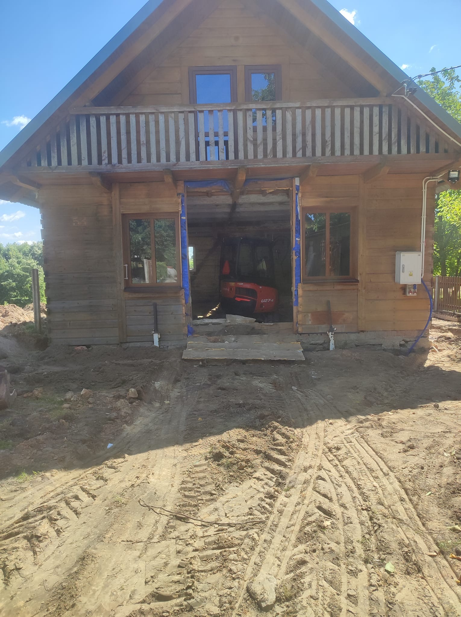 Mała czerwona koparka wewnątrz drewnianego domu w trakcie budowy; widoczne ślady opon na piaszczystym terenie przed budynkiem, sugerujące prace ziemne.