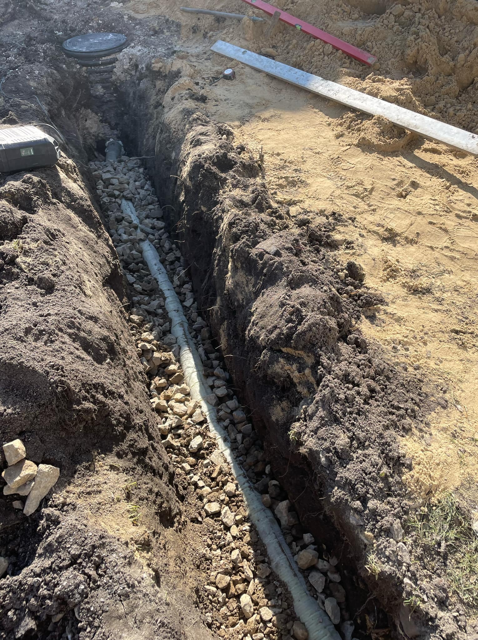Wykop pod instalację z ułożoną rurą, obsypaną kamieniami, widoczne narzędzia pomiarowe i studzienka kanalizacyjna w tle.