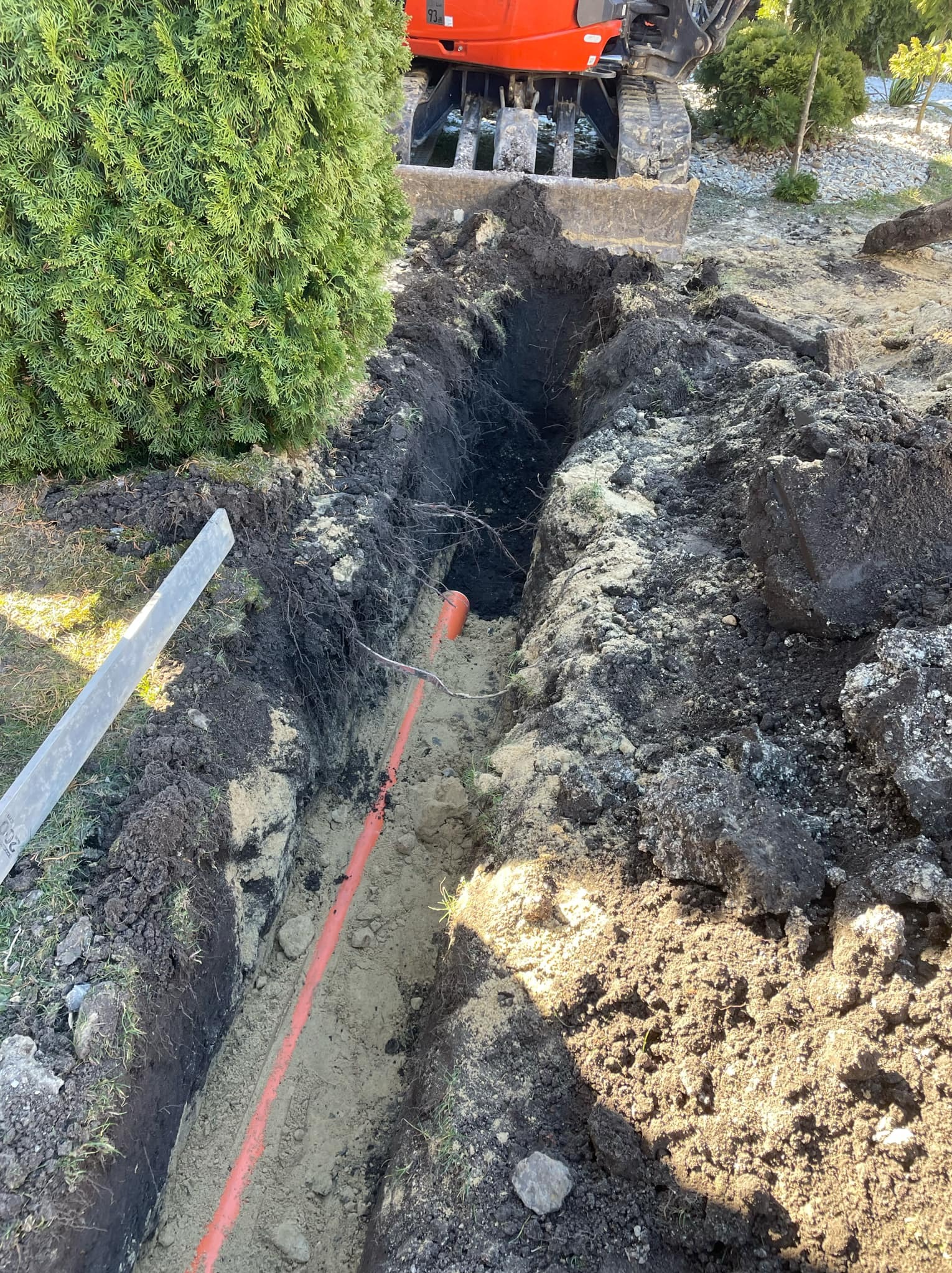 Wykopany rów z pomarańczową rurą na dnie, widoczna koparka z przodu, otoczenie to ziemia i roślinność.