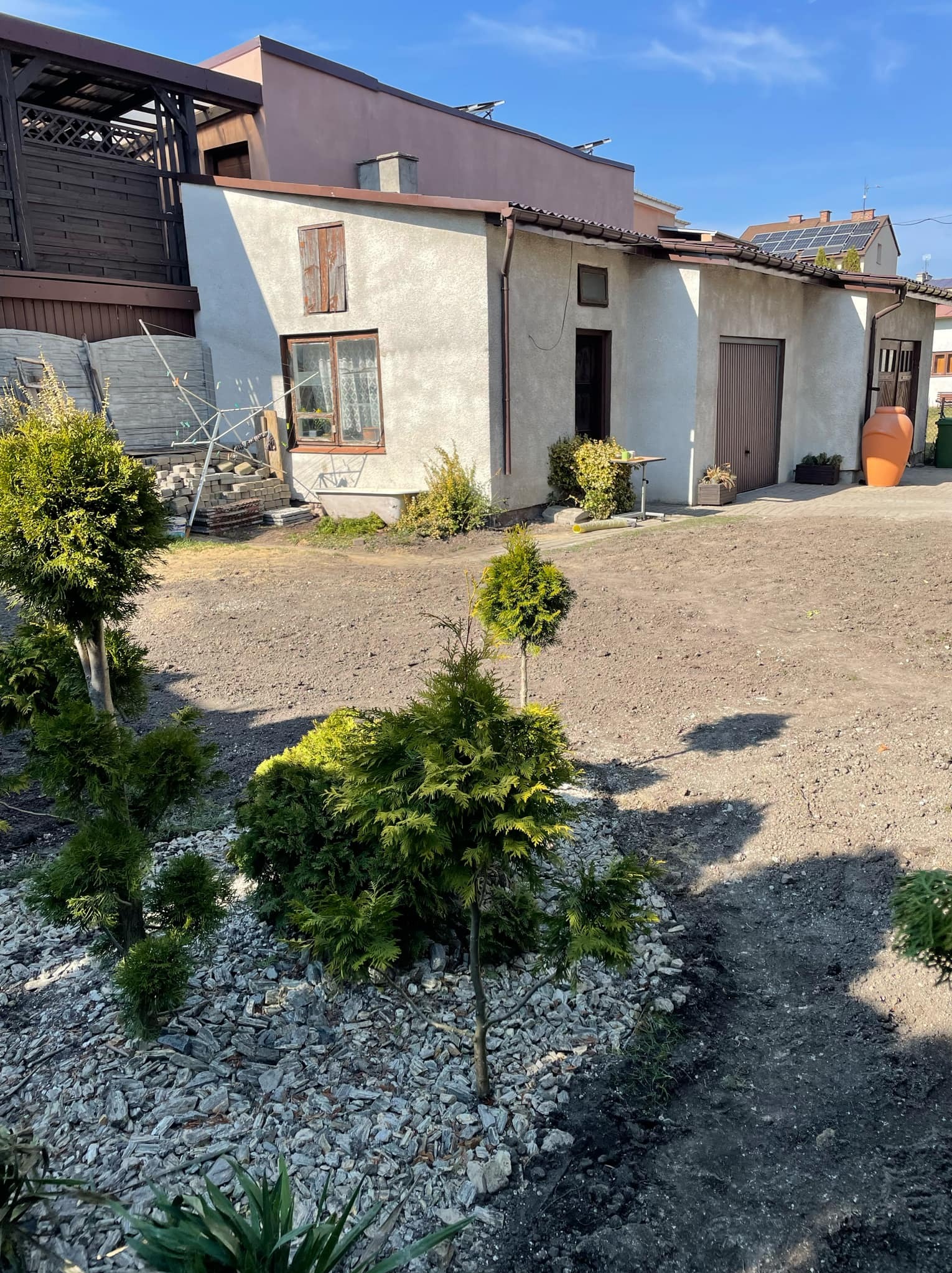 Przygotowany teren pod trawnik z nasadzeniami iglaków na tle domu z garażem i elementami drewnianej architektury ogrodowej.