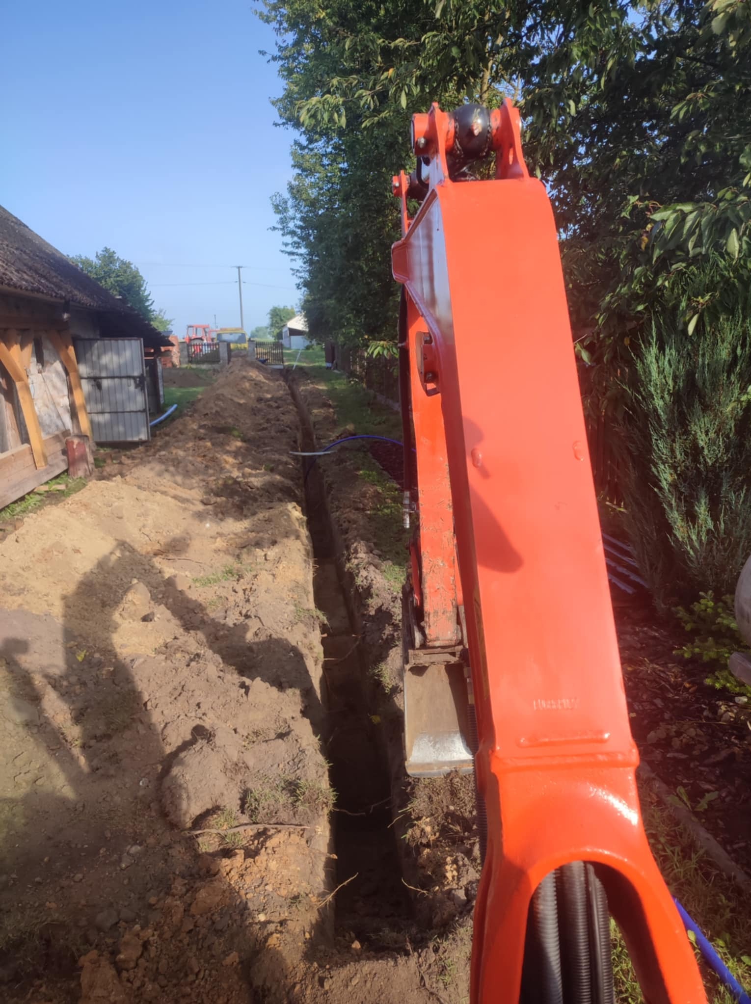 Pomarańczowa koparka w trakcie wykonywania głębokiego wykopu pod instalacje wzdłuż drewnianego budynku, widoczne rury instalacyjne w ziemi, w tle traktor i drzewa.