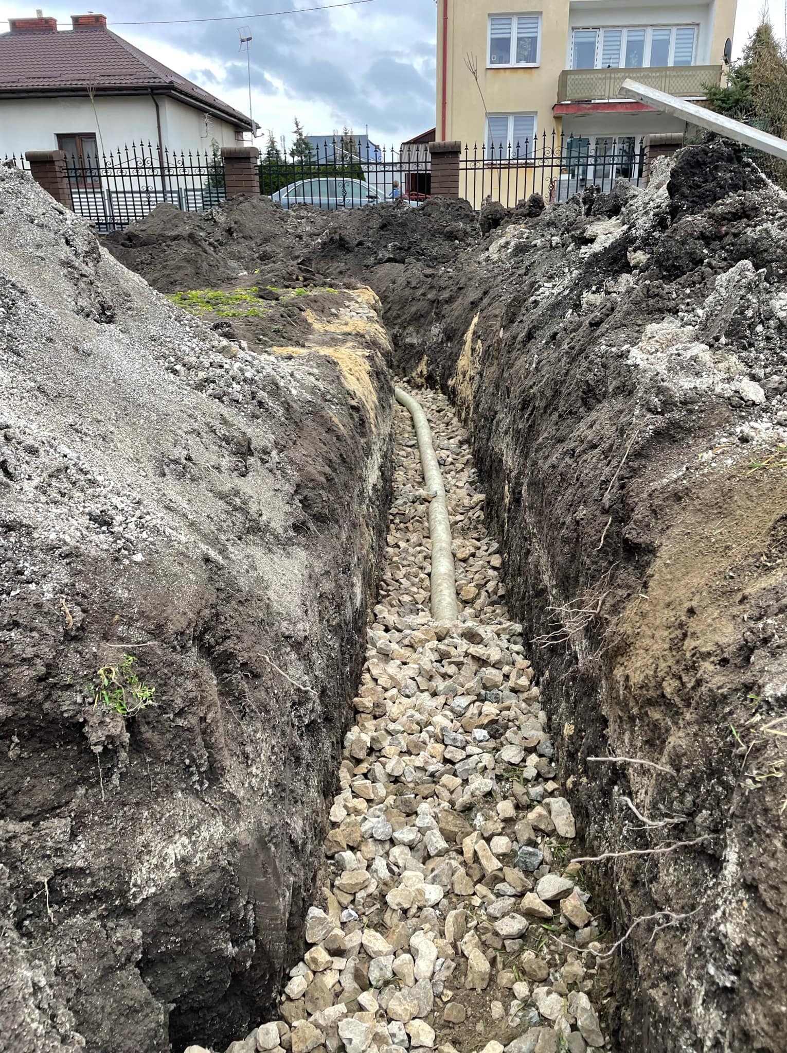 Wykopany wąski rów z ułożoną rurą na dnie, zasypany warstwą jasnych kamieni, z widoczną ziemią po bokach i fragmentem zabudowań w tle.