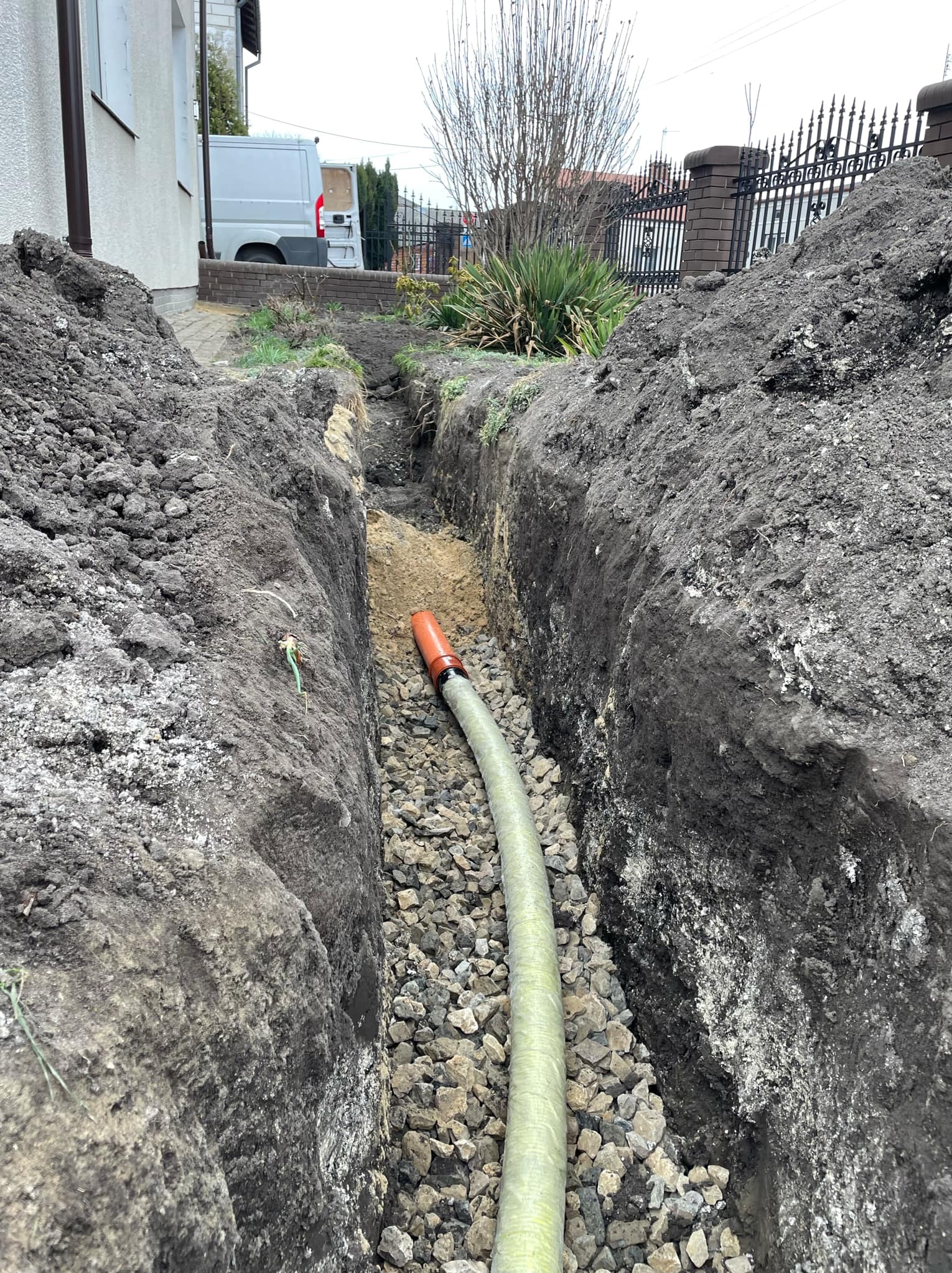 Wąski wykop w ziemi z ułożonym drenem na podsypce żwirowej, widoczny budynek w tle.