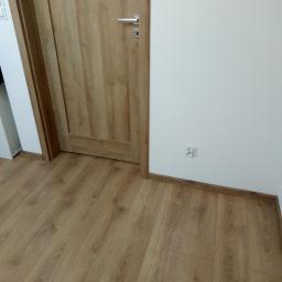 SAFEFORT Sp. z o.o. - Panel podłogowy w odcieniach dębu, drzwi wewnętrzne z klamką i gniazdko elektryczne przy białej ścianie.