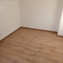 SAFEFORT Sp. z o.o. - Nowo ułożone panele podłogowe w jasnym odcieniu drewna w narożnym pokoju z pomalowanymi na biało ścianami; widoczny fragment wystającego przewodu elektrycznego.