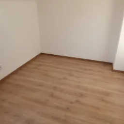 Nowo ułożone panele podłogowe w jasnym odcieniu drewna w narożnym pokoju z pomalowanymi na biało ścianami; widoczny fragment wystającego przewodu elektrycznego.