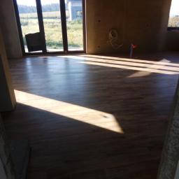 SAFEFORT Sp. z o.o. - Nowa podłoga z paneli ułożona w pokoju z dużym oknem tarasowym, widoczne promienie słoneczne wpadające do wnętrza, ściany w stanie surowym.