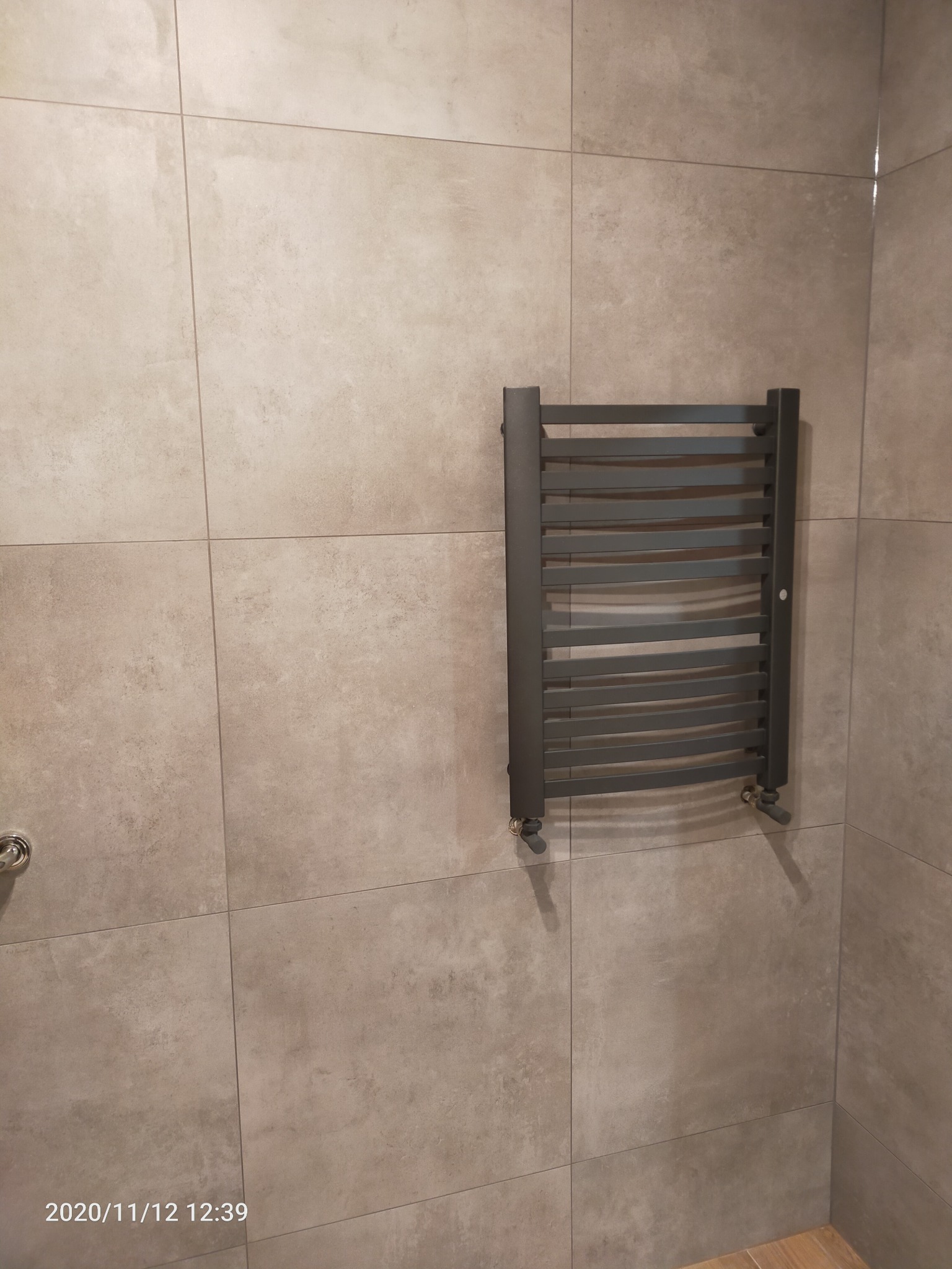 Szary grzejnik drabinkowy zamontowany na ścianie z płytek imitujących beton w łazience, widoczne zawory i fragment podłogi.