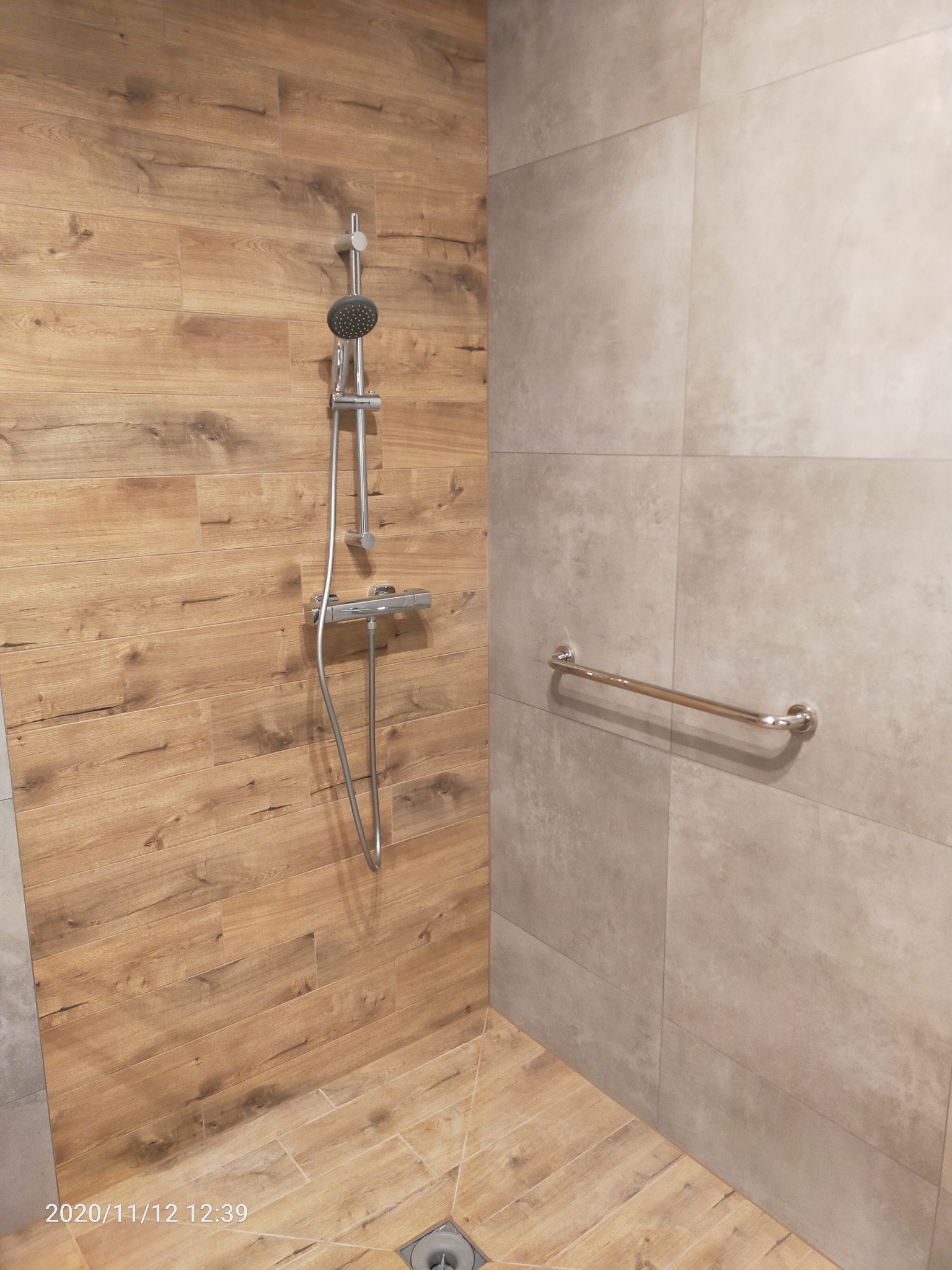 Nowoczesna kabina prysznicowa z armaturą chromowaną, ściany wyłożone płytkami imitującymi drewno i szarym gresem, uchwyt dla niepełnosprawnych.