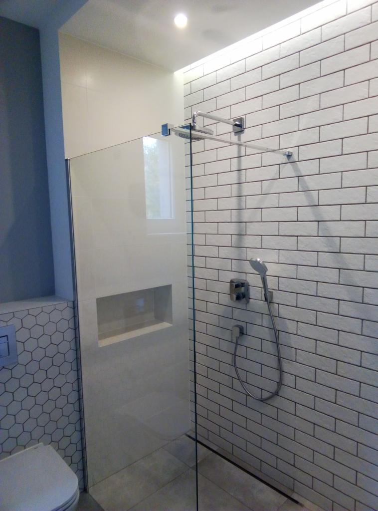 Nowoczesna kabina prysznicowa walk-in z przeszkloną ścianką, wbudowaną półką i białą cegłą na ścianie, uzupełniona chromowaną armaturą i heksagonalną mozaiką na sąsiedniej ścianie.