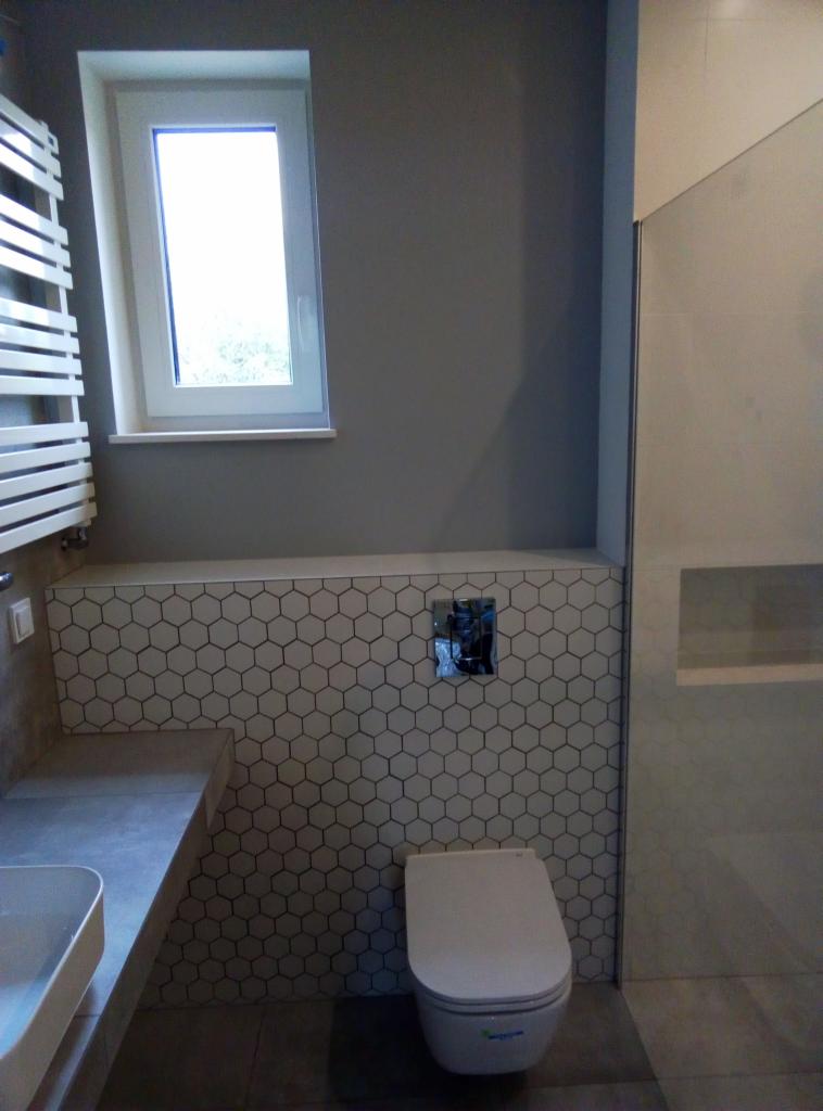 Nowoczesna łazienka z szarą ścianą, heksagonalnymi płytkami, podwieszaną toaletą i grzejnikiem drabinkowym przy oknie.