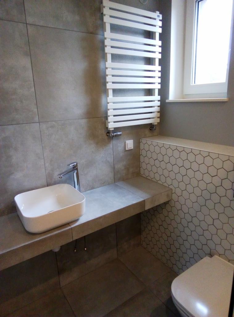 Nowoczesna łazienka z szarymi płytkami wielkoformatowymi, białą umywalką nablatową na betonowym blacie, grzejnikiem drabinkowym i toaletą z zabudową w heksagonalne wzory.