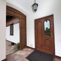 Wejście do domu w Woli Rzędzińskiej z brązowymi drzwiami zewnętrznymi z szybą, widoczny fragment elewacji i oświetlenie zewnętrzne.