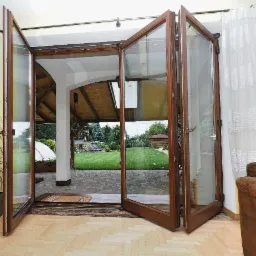 Rozłożone, brązowe drzwi harmonijkowe z widokiem na zielony ogród i altanę. W tle zasłona i roślina doniczkowa.