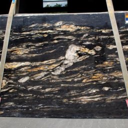 ANNAX - Czarny slab kamienny z wyraźnym, falistym wzorem w odcieniach beżu i bieli, zawieszony na pasach transportowych.