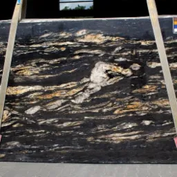 Czarny slab kamienny z wyraźnym, falistym wzorem w odcieniach beżu i bieli, zawieszony na pasach transportowych.