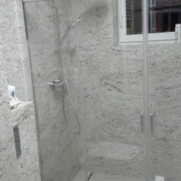 Wykończona łazienka z kabiną prysznicową, ściany i podłoga wyłożone płytkami imitującymi jasny kamień z delikatnym wzorem, widoczna toaleta i okno.