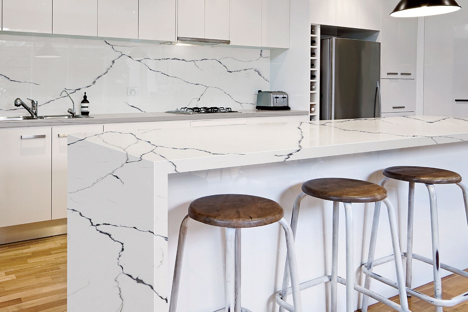 Nowoczesna kuchnia z białymi szafkami i blatem o wzorze imitującym marmur z czarnymi żyłami, uzupełniona o trzy drewniane stołki barowe z metalowymi nogami.