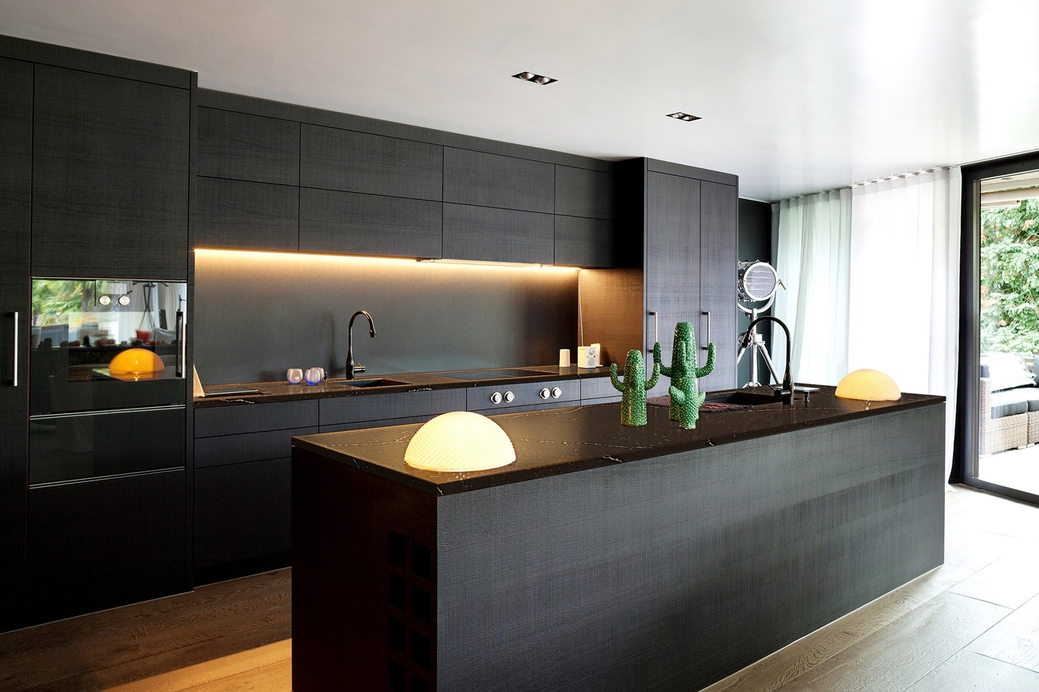 Nowoczesna kuchnia z czarnymi szafkami i blatem kamiennym o widocznej strukturze, oświetlona lampami dekoracyjnymi i podświetleniem LED pod szafkami wiszącymi, z dekoracyjnymi kaktusami na wyspie.