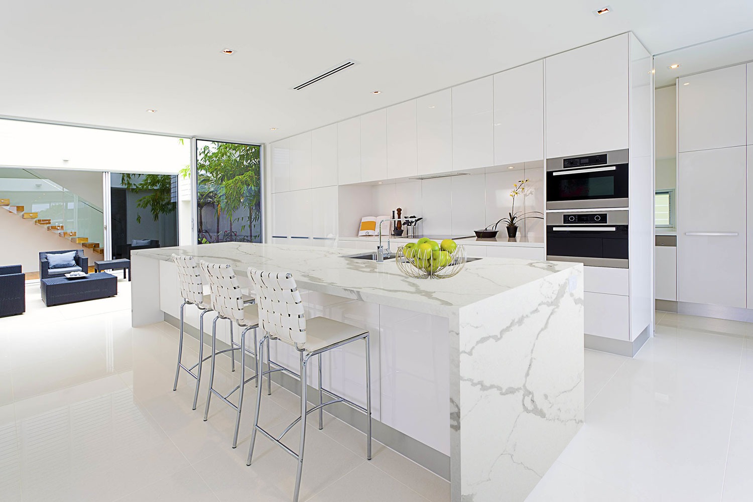 Nowoczesna, jasna kuchnia z białymi szafkami i wyspą z blatem o marmurowym wzorze, uzupełniona chromowanymi hokerami i koszem z zielonymi jabłkami.