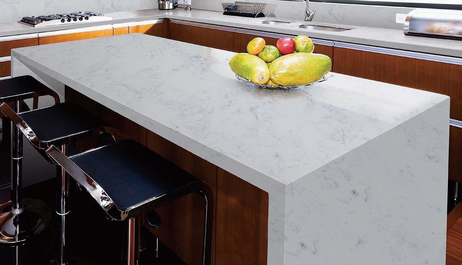 Szary blat kuchenny z delikatnym marmurkowym wzorem, z owocami w metalowej misie, na tle drewnianej zabudowy kuchennej i chromowanych stołków barowych.