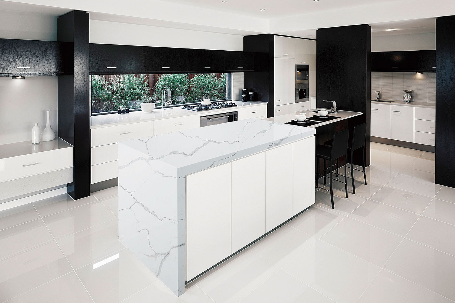 Nowoczesna kuchnia z białym marmurowym blatem wyspowym, czarne akcenty w szafkach i kolumnach, jasne płytki podłogowe, widok na zieleń przez okno.
