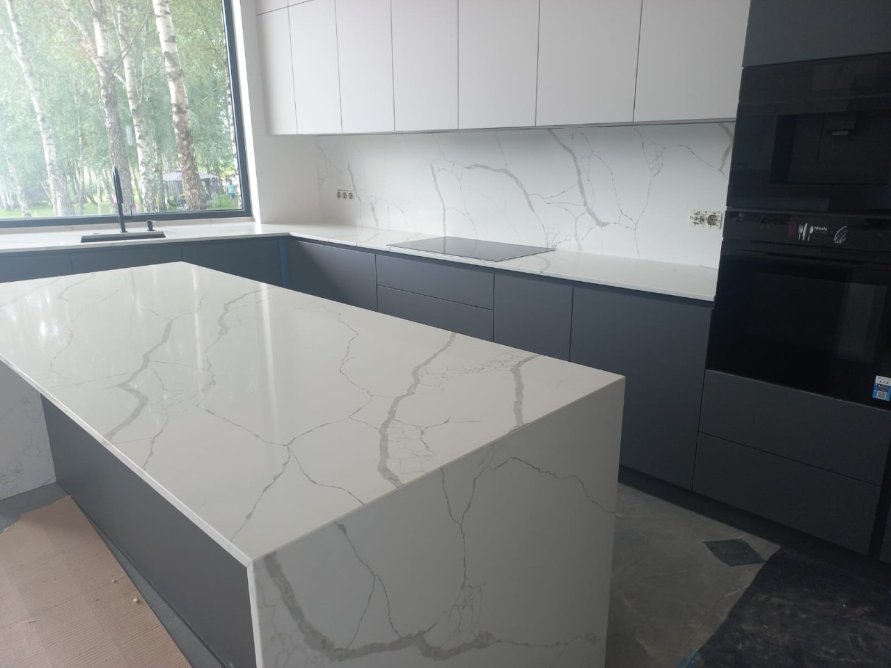 Nowoczesna kuchnia z szarymi szafkami i blatem z białego kamienia z wyraźnym żyłkowaniem, widok na okno z zielenią w tle.