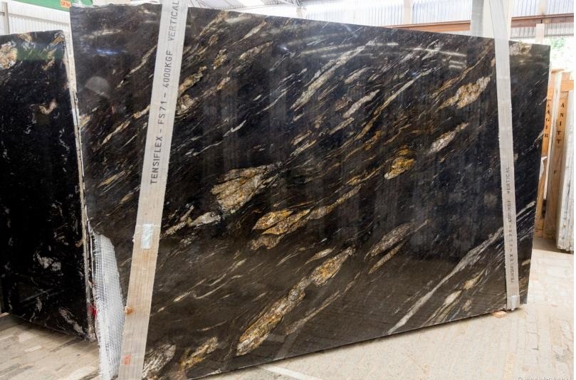 Dwa duże, zabezpieczone pasami transportowymi, polerowane płyty kamienne o czarnym tle z wyraźnymi, nieregularnymi, złotymi i białymi żyłami, widoczne w magazynie.