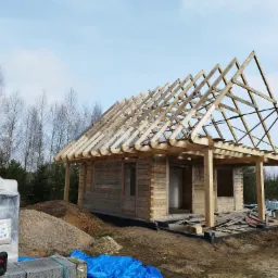 Surowy dom z bali w trakcie budowy, widoczna drewniana konstrukcja dachu, materiały budowlane i niebieska plandeka na ziemi.